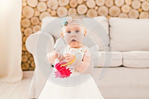 Little baby girl in white lavish dress standing in living room in house on sofa and wooden wall background and holding a