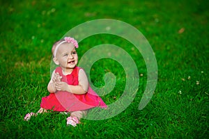 A little baby in a red dress sits on a grass