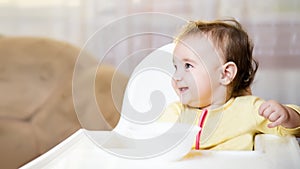 Little baby sitting in high chair and eating food indoors.. baby`s first feeding. 6 month old toddler holding spoon and plate and