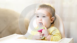 Little baby sitting in high chair and eating food indoors.. baby`s first feeding. 6 month old toddler with nibbler in the mouth