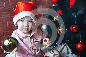 Happy baby in Santa s hat hiding behind a ball against Christmas tree with decorations. Ball in focuse. Baby unfocused.