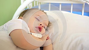 Little baby sleeping with thermometer under his arm in hospital ward on white bed. Sick child measures temperature in