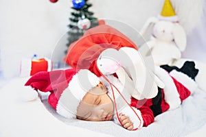 Little baby wearing Santa Claus costume sleep on white fur carpet with red gift bag and doll