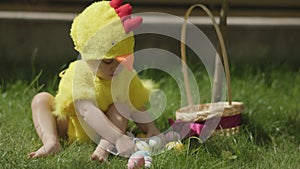 little baby in yellow chick costume pick up easter eggs from green grass