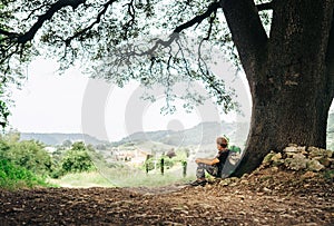 Little backpacker traveler rests under big tree on country road