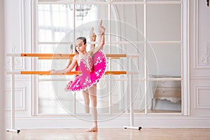 Little ballerina in a bright pink tutu is engaged in a ballet barre in front of a mirror in a beautiful white hall