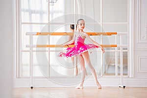 Little ballerina in a bright pink tutu is engaged in a ballet barre in front of a mirror in a beautiful white hall