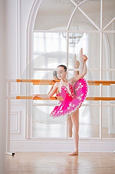 Little ballerina in a bright pink tutu is engaged in a ballet barre in front of a mirror in a beautiful white hall