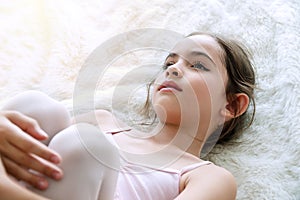 Little ballet girl brown long hair lying on white fur rug while looking at camera . Top view lovely kid lying down