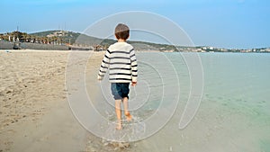 Little barefoot boy running on sandy sea beach and splashing water. Concept of kids traveling, joy, fun, happiness, holiday,