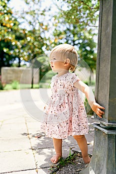 Little barefoot girl in a dress stands on a tile in the park near the pillar