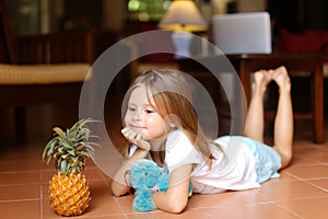 Little barefoot girl lying on floor with pineapple and toy.