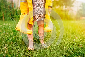 Little barefoot girl in yellow raincoat standing in the grass and holding yellow rubber boots in her hands. Summertime