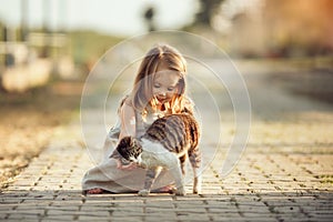 A little barefooted girl in a gray linen dress is stroking a cat. Sunny summer evening in the village. Countryside