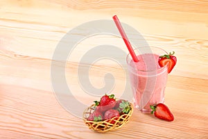 Little basket full of strawberries is standing on a desk next to transparent glass of delish coral smoothie with a red straw.