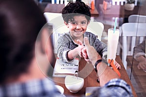 Little beaming dark-haired son looking at his father sitting in cafe together