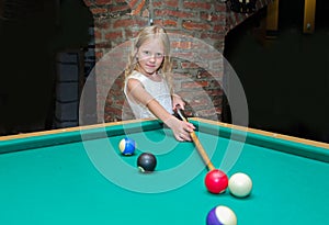 Little beautiful blond girl learns to play billiards, pool, snooker, Russian pyramid in kid`s club