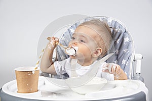 Little beautiful child eating a wooden spoon of yogurt from a plate and a paper cup with a straw with milk on a high gray chair on