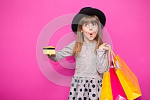 Little beautiful child girl in black hat holding shopping bags and credit card isolated on pink