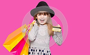 Little beautiful child girl in black hat holding shopping bags and credit card isolated on pink