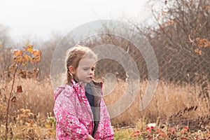 Little beautiful cute sad girl in a jacket with blond hair in pigtails stands on an autumn background