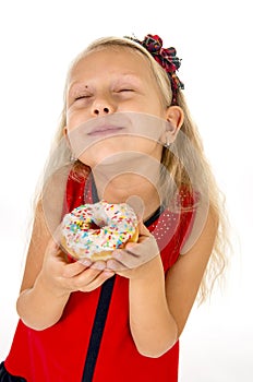 Little beautiful female child with long blonde hair and red dress eating sugar donut with toppings delighted and happy