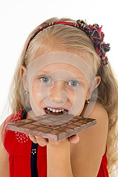 Little beautiful female child in red dress holding happy delicious chocolate bar in her hands eating delighted