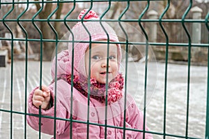 Little beautiful girl baby behind the fence, grid locked in a cap and a jacket with sad emotion on his face