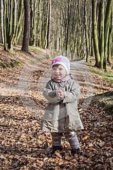 Little beautiful girl in a baby raincoat, hat and scarf is played in spring forest dry leaf litter throwing their smiles in a good