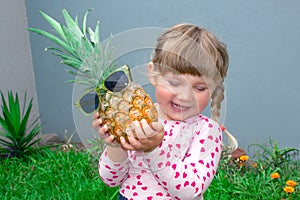 The little beautiful girl cheerfully smiles, laughs and holds in hands pineapple in glasses. In the garden outdoors.