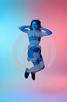Little beautiful girl, child listening to music in headphones and jumping over gradient blue pink studio background in