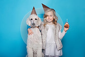 Little beautiful girl with dog celebrate birthday. Friendship. Love. Cake with candle. Studio portrait over blue background