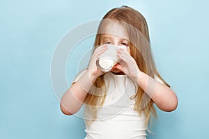 Little beautiful girl drinking milk in glass on a blue background