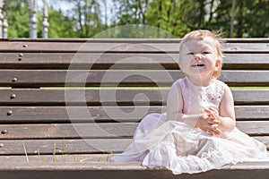 Little Beautiful Girl in Elegant Dress Sits on a Park Bench and Laughs. Summer Concept