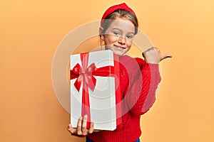 Little beautiful girl holding gift pointing thumb up to the side smiling happy with open mouth