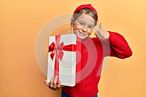 Little beautiful girl holding gift smiling happy and positive, thumb up doing excellent and approval sign