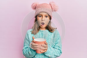 Little beautiful girl holding pink mug wearing cute wool hat afraid and shocked with surprise and amazed expression, fear and