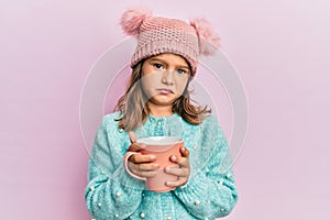 Little beautiful girl holding pink mug wearing cute wool hat clueless and confused expression