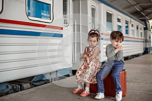 Little beautiful girl in retro dress says goodbye at the station with a little boy in vintage clothes with retro suitcase