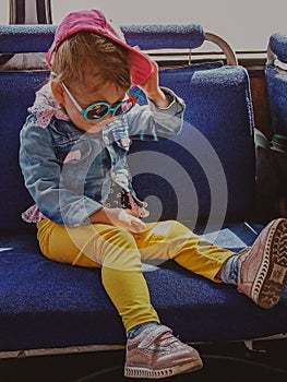 Little beautiful girl sits in a chair on public transport in yellow pants and blue jacket and glasses