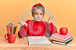 Little beautiful girl sitting on classroom desk clueless and confused expression with arms and hands raised