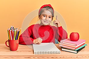 Little beautiful girl sitting on classroom desk pointing finger up with successful idea