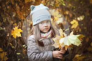 Little beautiful girl stands with a bouquet of maple orange leaves in the autumn park. They are looking at the camera. Dressed in