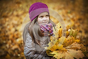 Little beautiful girl stands with a bouquet of maple orange leaves in the autumn park. She looks away and smiles. Dressed in a