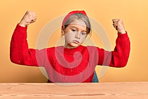 Little beautiful girl wearing casual clothes sitting on the table showing arms muscles smiling proud