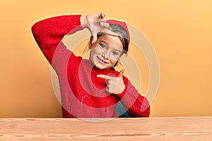 Little beautiful girl wearing casual clothes sitting on the table smiling making frame with hands and fingers with happy face