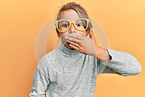 Little beautiful girl wearing casual clothes and yellow glasses covering mouth with hand, shocked and afraid for mistake