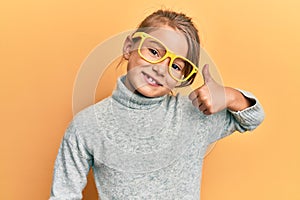 Little beautiful girl wearing casual clothes and yellow glasses smiling happy and positive, thumb up doing excellent and approval