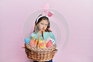 Little beautiful girl wearing cute easter bunny ears holding wicker basket with colored eggs clueless and confused expression