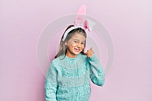 Little beautiful girl wearing cute easter bunny ears smiling with happy face looking and pointing to the side with thumb up